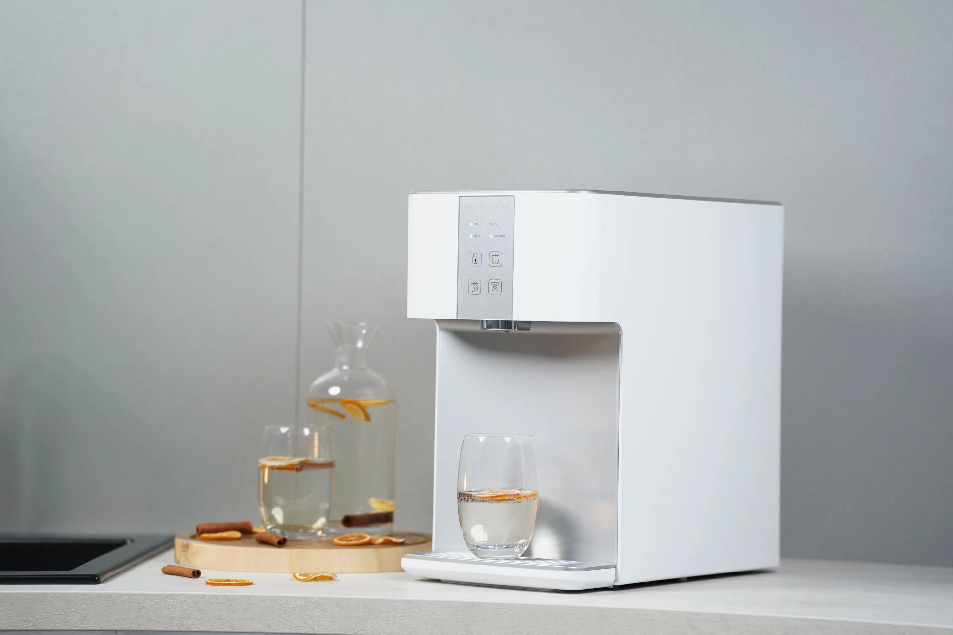 Ein moderner Wasserspender, die Sparkling Pro der Marke Aqua Global auf einer Küchenarbeitsfläche, der eine Glas-Teekanne mit heißem Wasser füllt. Daneben sind Zimtstangen, getrocknete Orangenscheiben und Gebäck dekorativ arrangiert.